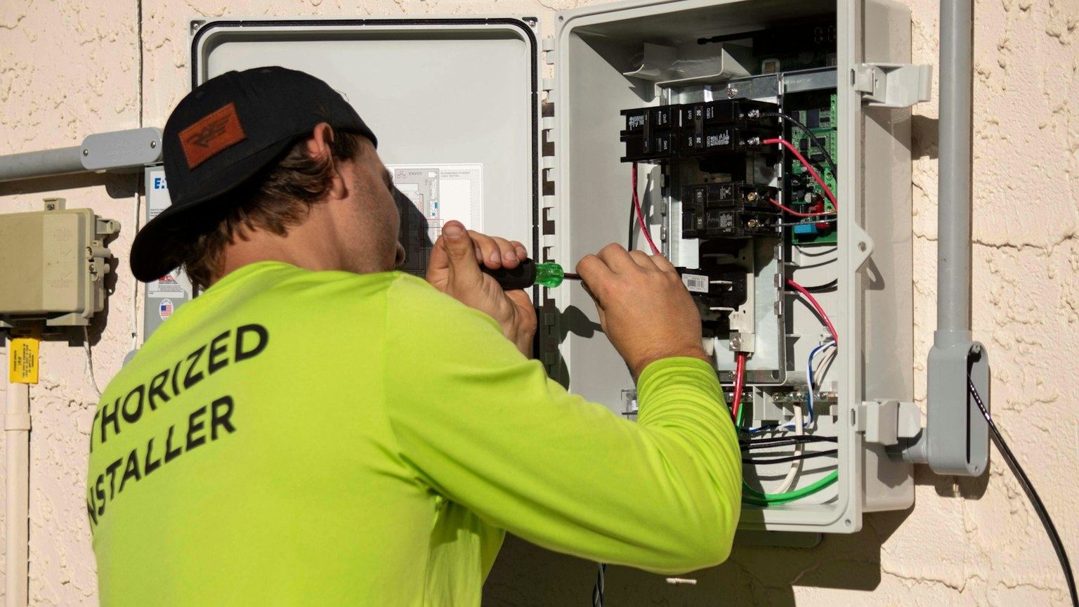 An electrician wiring a home energy panel mounted on an exterior stucco wall
